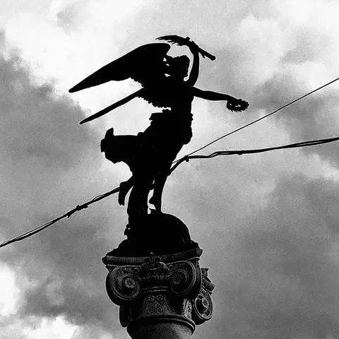Silhouette di una statua alata con lama in mano in cima a una colonna decorata, contro un cielo nuvoloso, in bianco e nero