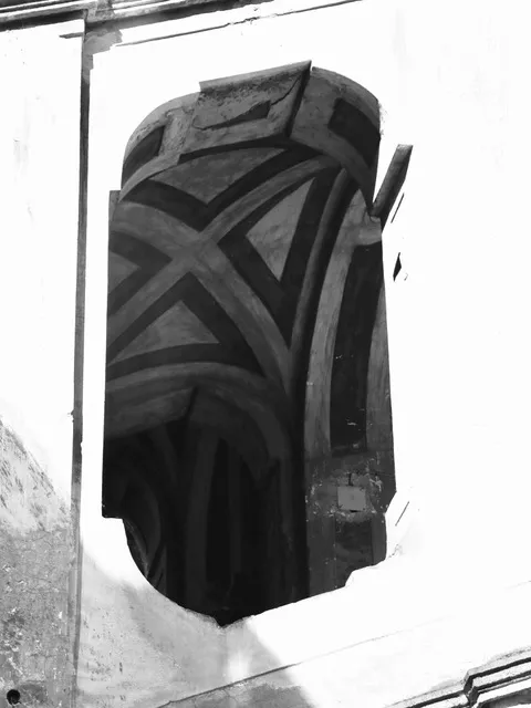 Dettaglio architettonico scuro dalla forma arcuata con bande incrociate, incassato in una parete chiara, in bianco e nero
