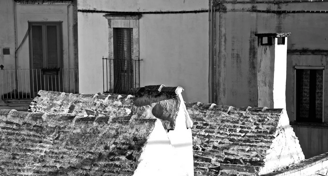 Tetti in pietra e tegole di antiche case, con facciate imbiancate, balconi e comignoli sullo sfondo, in bianco e nero