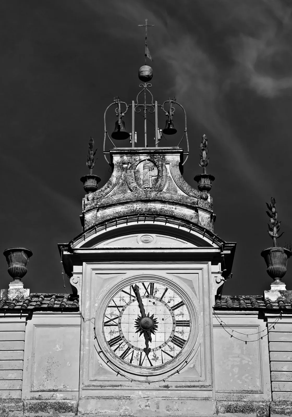 Orologio con numeri romani e motivo a sole in cima a un edificio, due campane sopra, cielo scuro, in bianco e nero