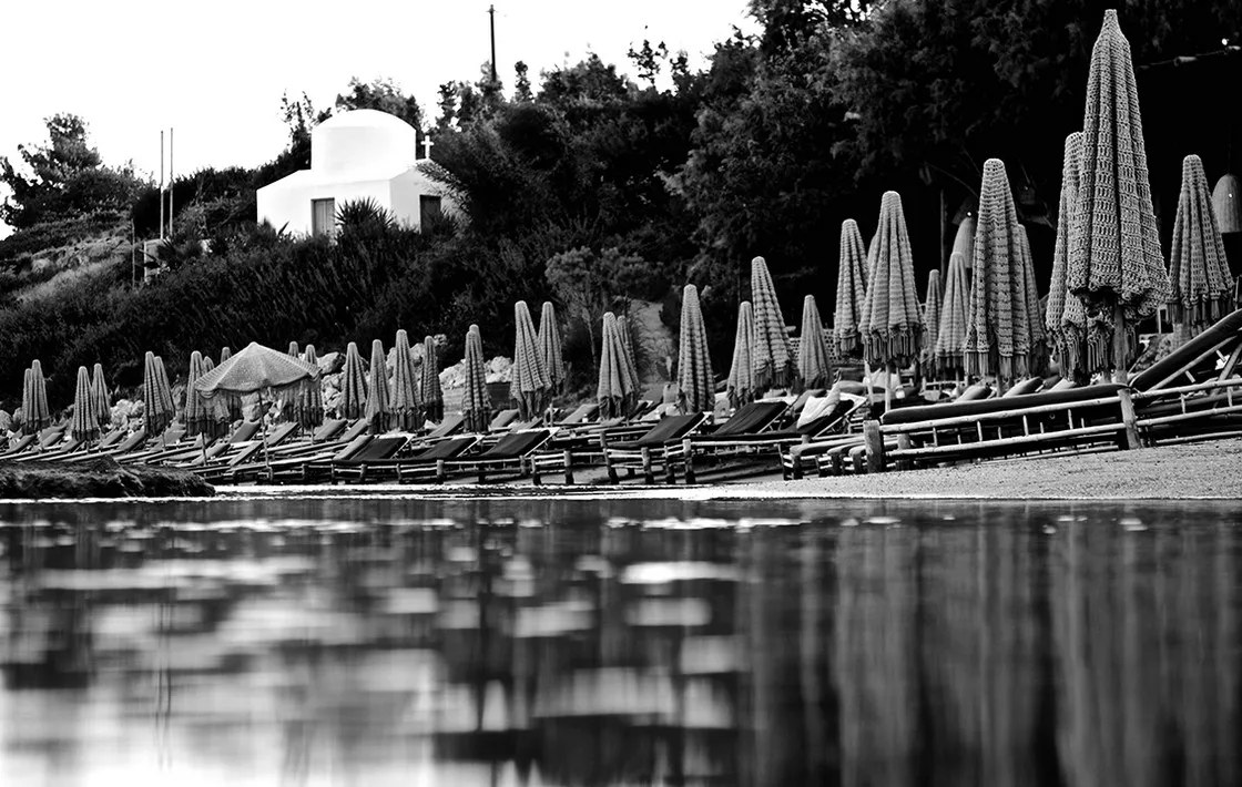 Fila di ombrelloni di paglia chiusi e lettini lungo una spiaggia, cappella bianca su un'altura, riflessi nell'acqua, in bianco e nero