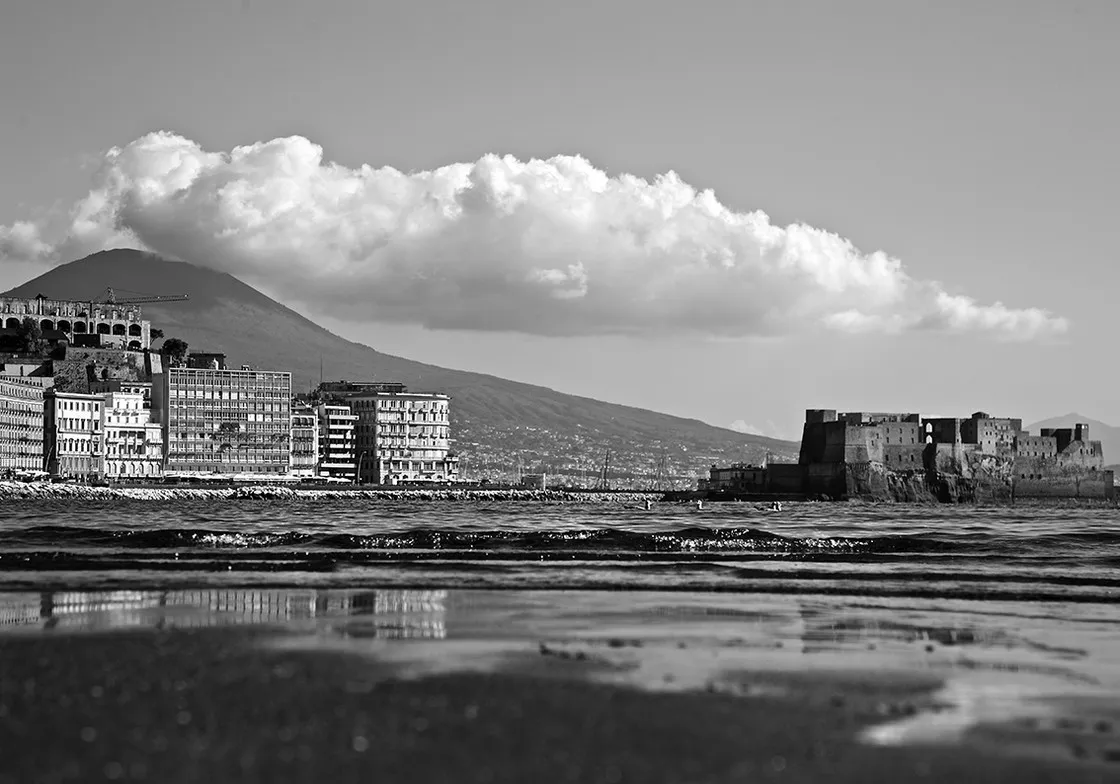 Veduta di un lungomare con palazzi, un castello sull'acqua e un vulcano coronato da una nuvola, in bianco e nero