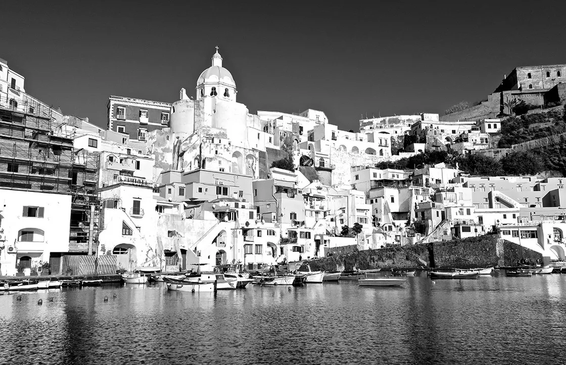 Case addossate sulla collina di un borgo marinaro affacciato sul porto, con barche ormeggiate, in bianco e nero