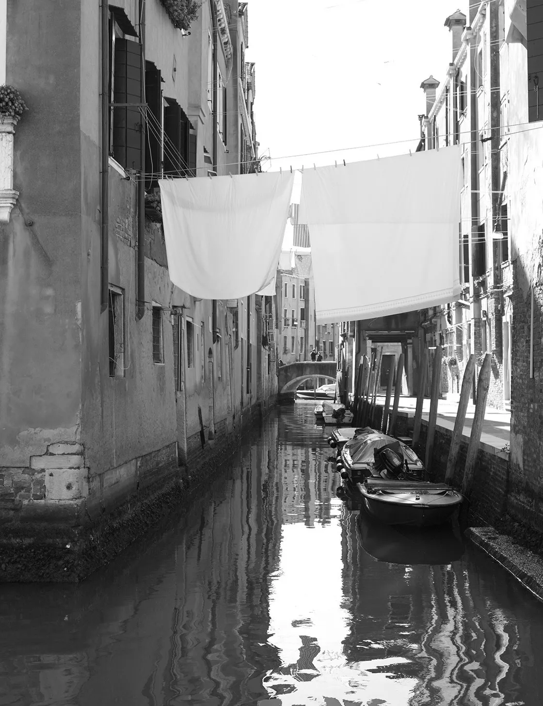 Stretto canale con lenzuola bianche stese ad asciugare tra i palazzi e barche ormeggiate, in bianco e nero