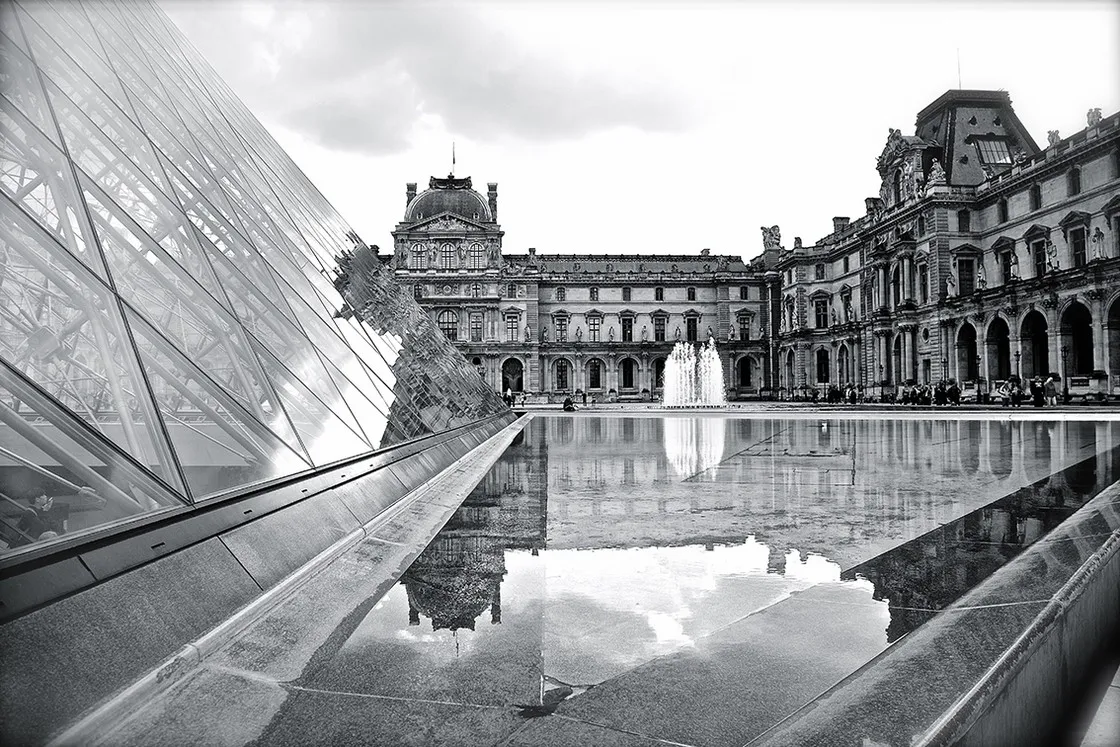 Piramide di vetro e facciata di un grande palazzo storico si riflettono in una vasca con getto d'acqua, in bianco e nero