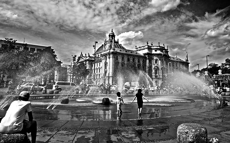 Bambini giocano tra gli spruzzi di una fontana in una piazza, grande palazzo storico con cupola sullo sfondo, in bianco e nero