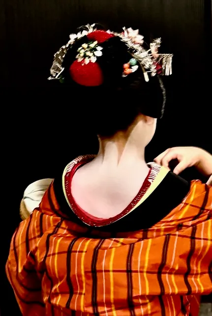 Maiko vista di spalle, con acconciatura ornata di fiori e kimono arancione a quadri