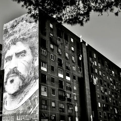 Il murale di Jorit con il volto di Maradona su una torre di edilizia popolare, tra i balconi e un ramo di pino, in bianco e nero