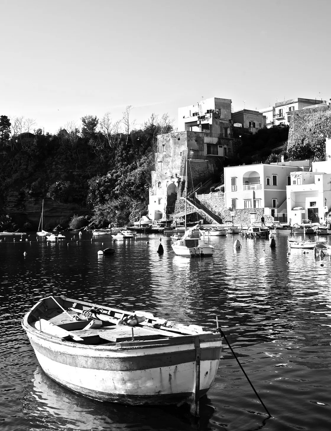 Barca da pesca in legno ormeggiata in una piccola insenatura, con case sulla collina alle spalle, in bianco e nero