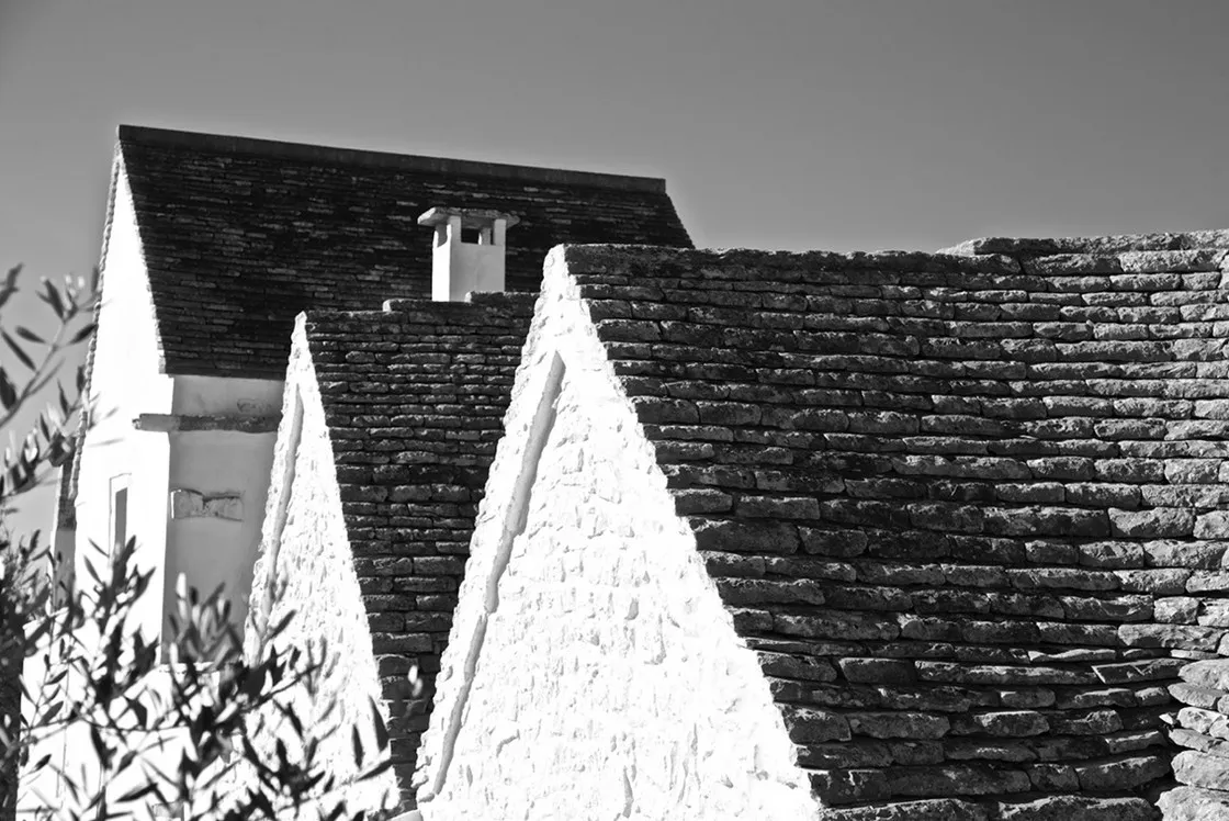 Tetti conici in pietra a secco e una casa imbiancata, tra rami d'ulivo, sotto il cielo, in bianco e nero