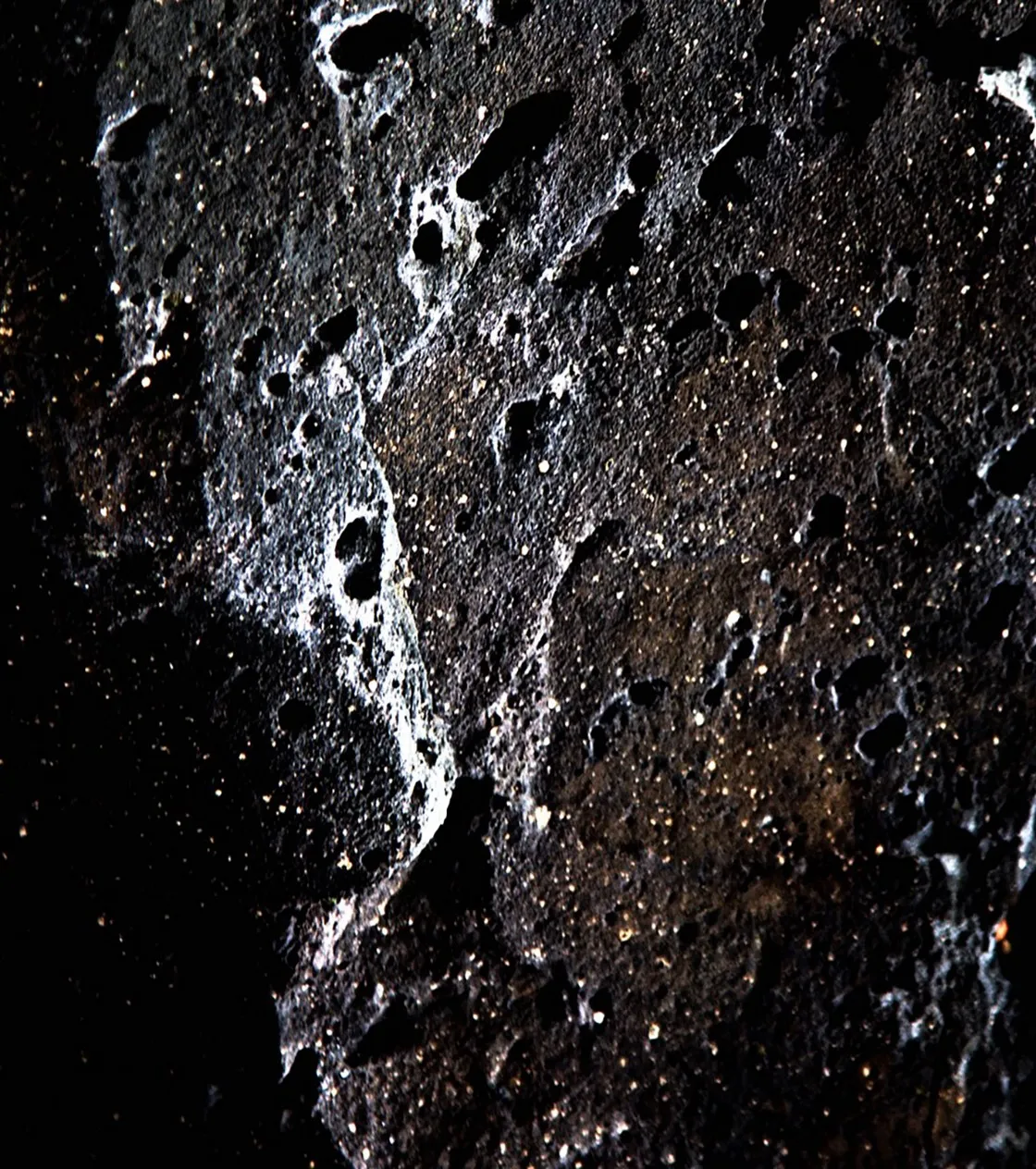 Superficie scura e porosa di roccia vulcanica costellata di cavità e riflessi luminosi