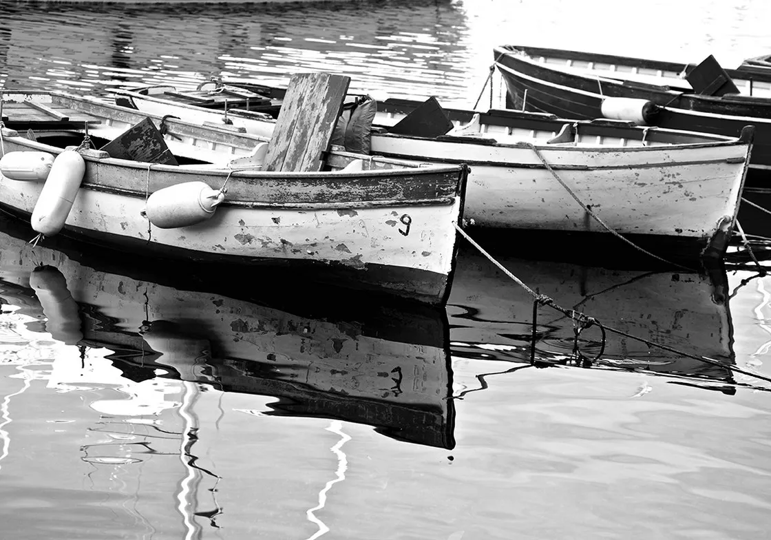 Barche da pesca in legno dalla vernice scrostata, ormeggiate su acqua calma che le riflette, in bianco e nero