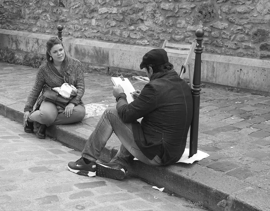 Ritrattista di strada seduto sul cordolo disegna una giovane donna seduta di fronte, su pavé, in bianco e nero