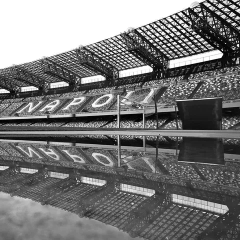 La scritta NAPOLI sugli spalti dello stadio, riflessa capovolta in una pozza d'acqua a bordo campo, in bianco e nero