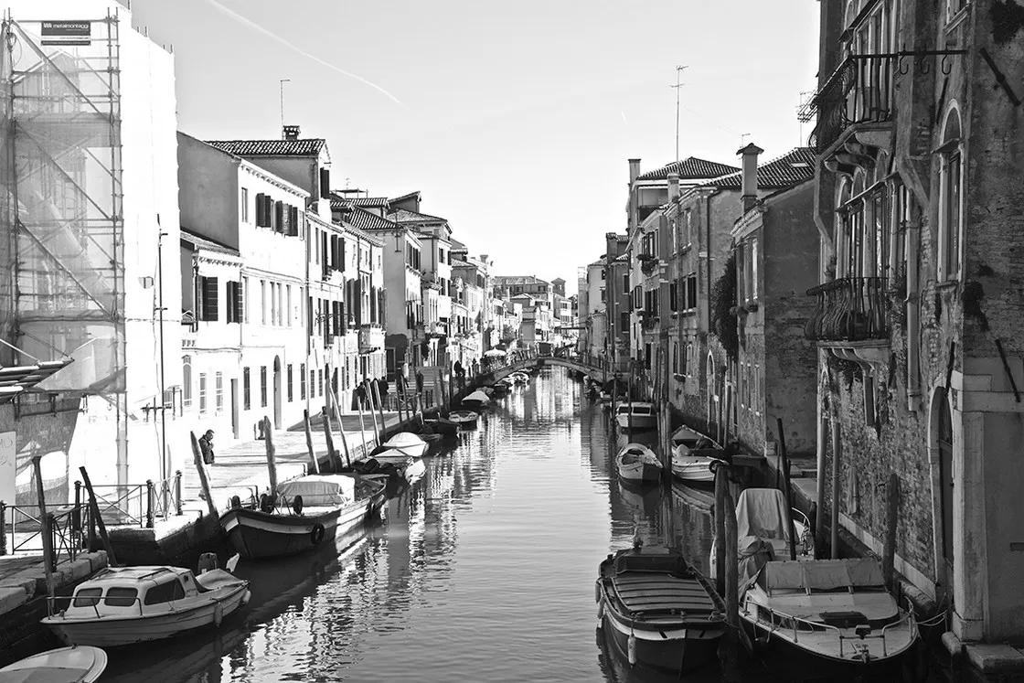 Canale con palazzi su entrambi i lati, barche ormeggiate e un ponte in fondo, in bianco e nero