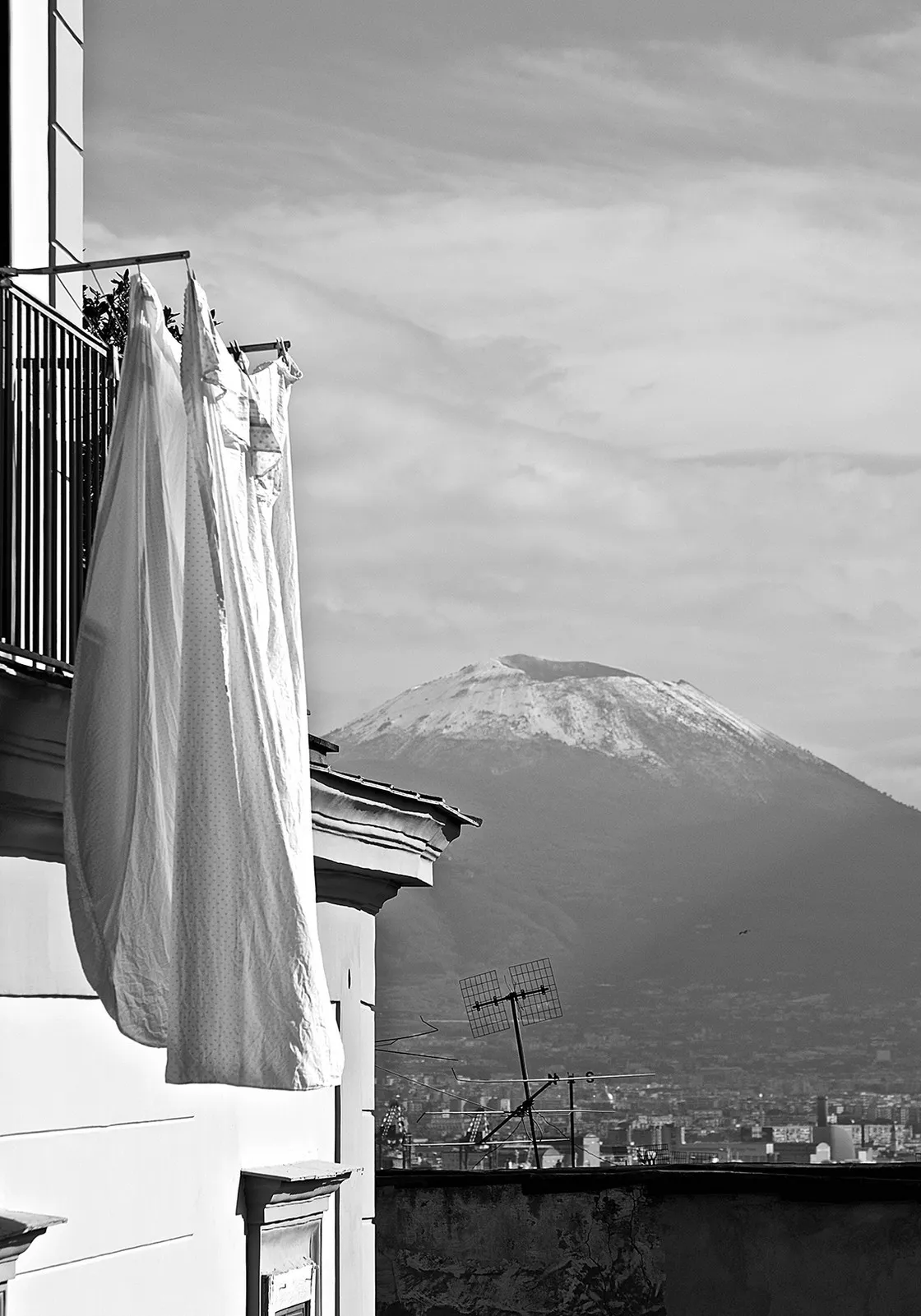 Lenzuola bianche stese al balcone e, sullo sfondo, un monte innevato che domina i tetti della città, in bianco e nero