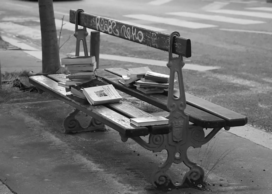 Panchina di legno con fianchi in ghisa coperta di libri impilati, lungo un marciapiede, in bianco e nero