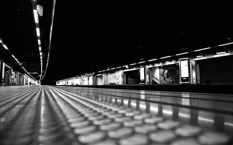 Banchina deserta di una stazione metropolitana ripresa dal basso con le luci in lontananza, in bianco e nero