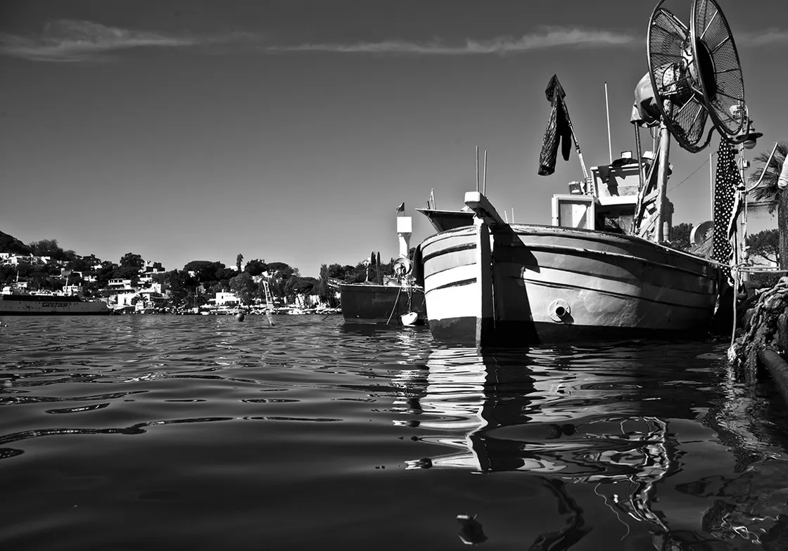 Barche da pesca ormeggiate viste dal pelo dell'acqua, con il borgo sulla costa sullo sfondo, in bianco e nero