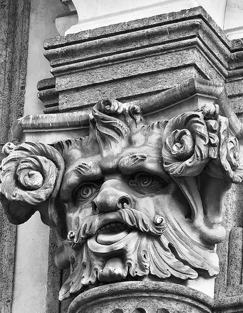 Mascherone in pietra scolpito con volto barbuto e volute fogliate, sotto una cornice modanata, in bianco e nero