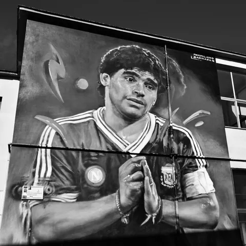 Murale con Maradona in maglia dell'Argentina, le mani giunte e lo sguardo commosso, dipinto su una parete tra le finestre, in bianco e nero
