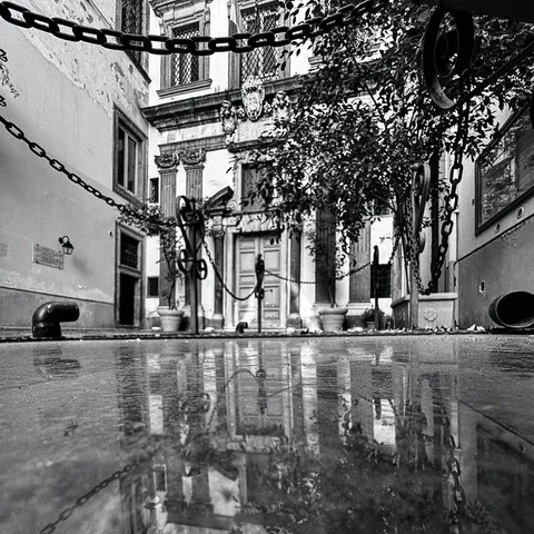 Cortile monumentale con catene di dissuasione in primo piano e un portale antico riflesso nel basolato bagnato, in bianco e nero