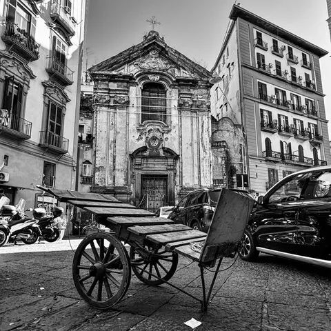 Un carretto di legno a due ruote fermo davanti alla facciata barocca di una chiesa del centro storico, in bianco e nero