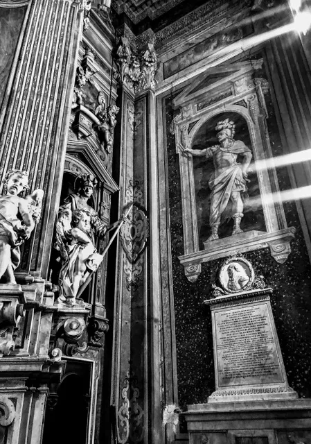 Interno barocco con statue in marmo, una nicchia con figura in armatura e una lapide incisa, attraversato da lame di luce, in bianco e nero