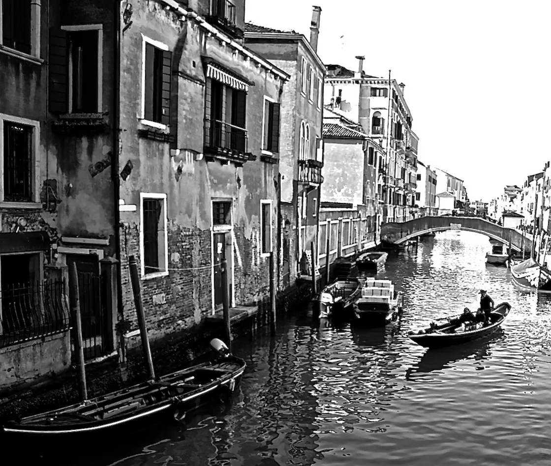 Una persona voga su una piccola barca lungo un canale tra palazzi scrostati, verso un ponte, in bianco e nero