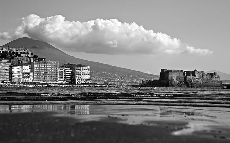 Palazzi sul lungomare e un castello sul mare con il Vesuvio e una grande nuvola sullo sfondo, in bianco e nero