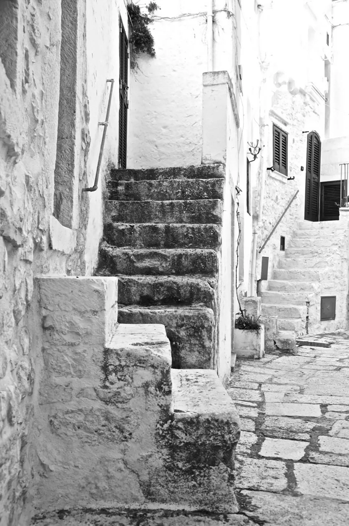 Stretta scalinata in pietra tra case imbiancate, con vicolo lastricato e persiane, in bianco e nero