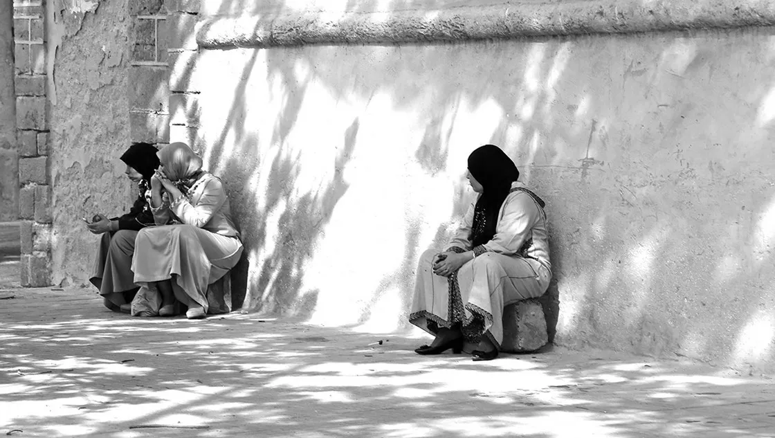 Tre donne con velo sedute contro un muro chiaro, ombre di fogliame sulla parete, in bianco e nero