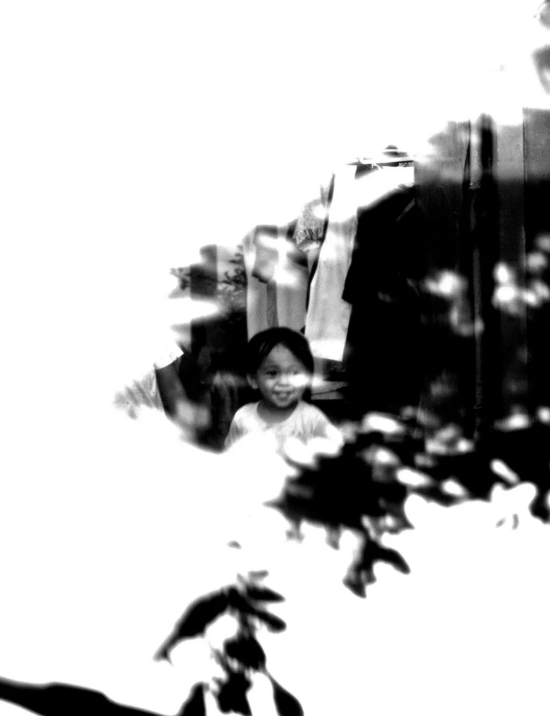 Bambino sorridente appena visibile tra panni stesi e foglie, su uno sfondo bianco e sovraesposto, in bianco e nero