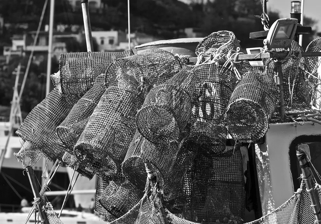 Nasse da pesca in rete accatastate su un peschereccio, con case sulla collina alle spalle, in bianco e nero