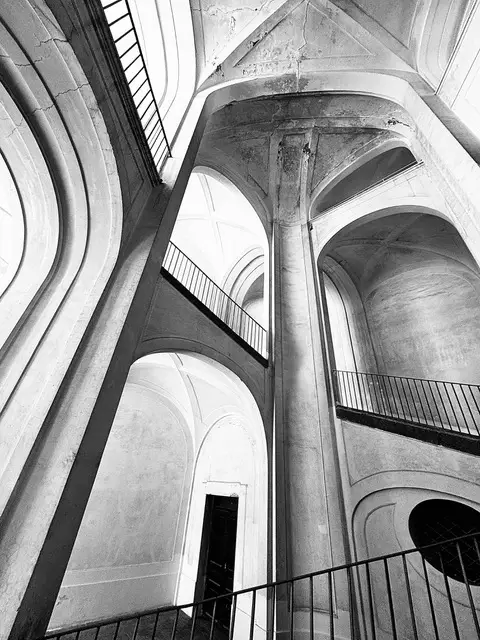 Scalone monumentale visto dal basso tra arcate, volte e una grande colonna centrale, con ringhiere in ferro, in bianco e nero