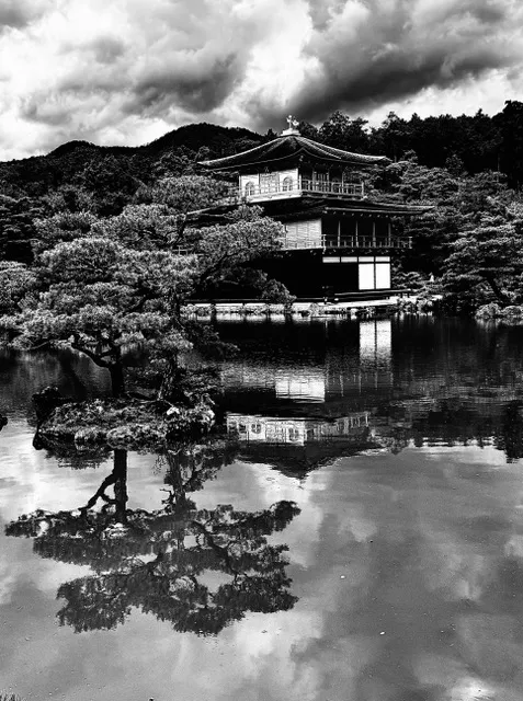 Padiglione tradizionale a più piani riflesso sull'acqua di un laghetto, tra alberi e cielo nuvoloso, in bianco e nero