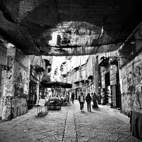 Vicolo del centro storico coperto da una rete ombreggiante, con banchi di mercato e passanti sul basolato, in bianco e nero