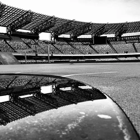 Gli spalti e le coperture dello stadio riflessi su una superficie lucida a bordo campo, in bianco e nero