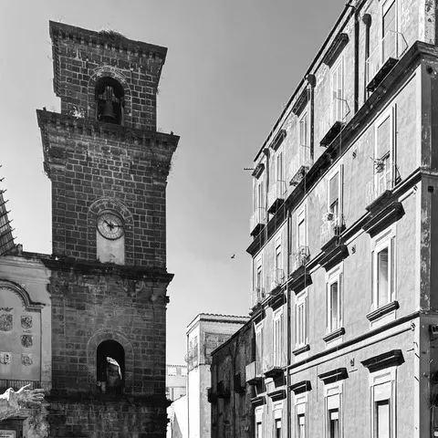 Campanile in piperno con orologio e cella campanaria accanto a un palazzo dalle file di balconi, in bianco e nero
