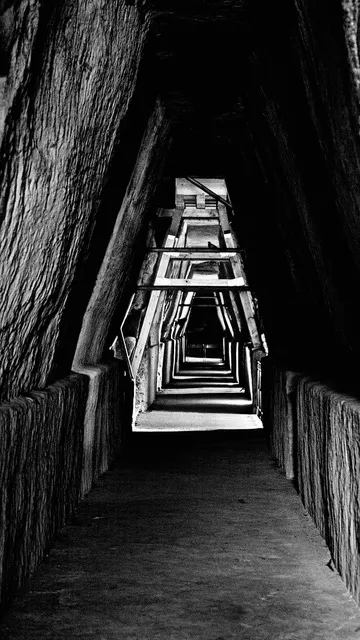 Stretto tunnel triangolare con travi di legno che si susseguono verso la luce in fondo, in bianco e nero