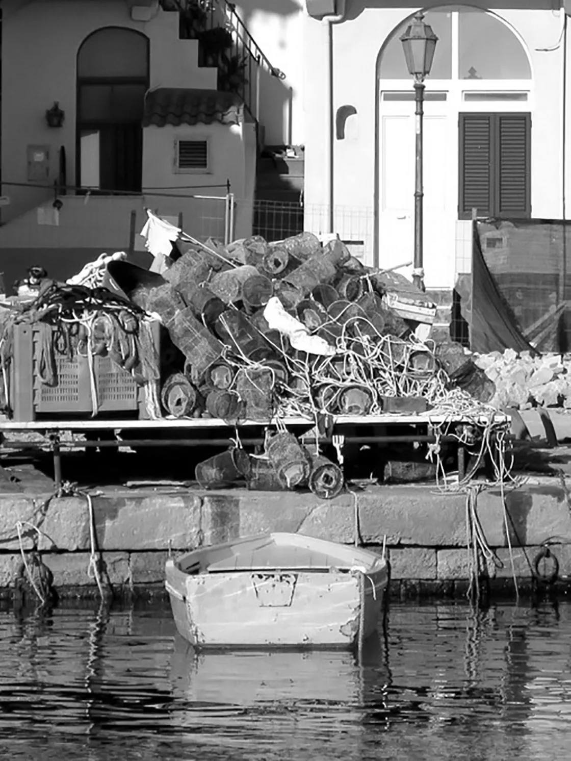 Nasse e reti da pesca ammassate sulla banchina accanto a un lampione, con una barca a remi, in bianco e nero