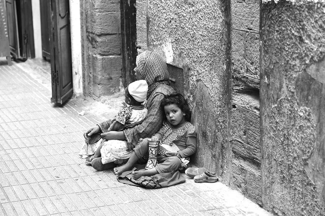 Una donna con foulard e due bambini piccoli seduti a terra contro un muro di pietra in strada, in bianco e nero