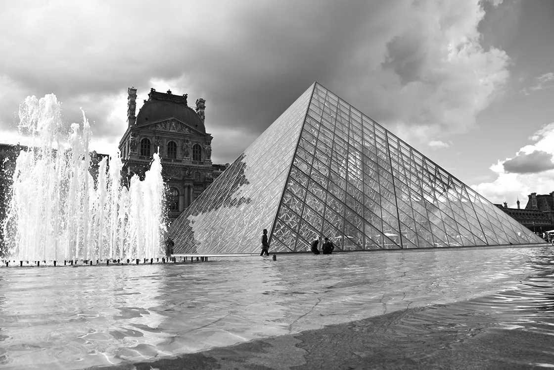 Piramide di vetro accanto a un palazzo storico, fontane zampillanti e cielo nuvoloso, in bianco e nero