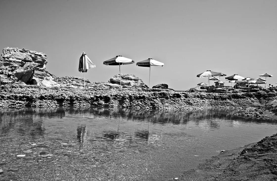 Ombrelloni aperti su una costa rocciosa si riflettono nell'acqua bassa e calma, in bianco e nero