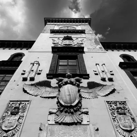 Facciata con torre dell'orologio e una grande aquila scolpita ad ali spiegate tra ghirlande e stemmi, ripresa dal basso, in bianco e nero