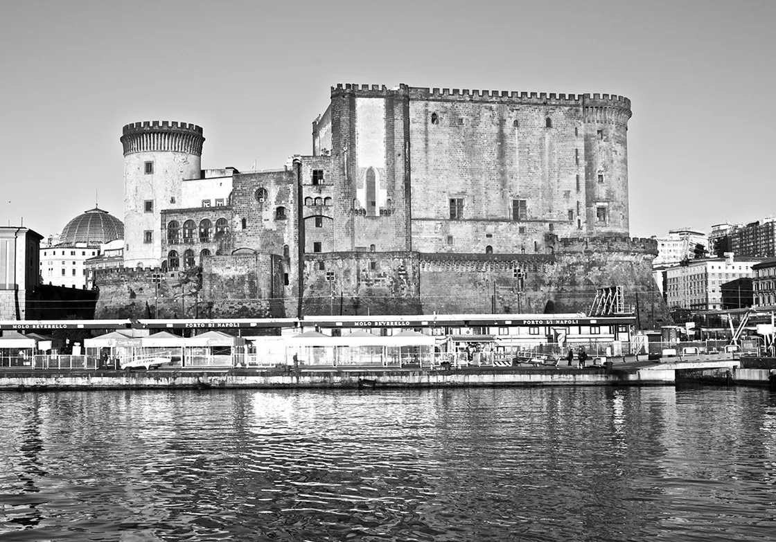 Imponente castello medievale con torri cilindriche e merlature visto dal mare, con il porto in primo piano, in bianco e nero