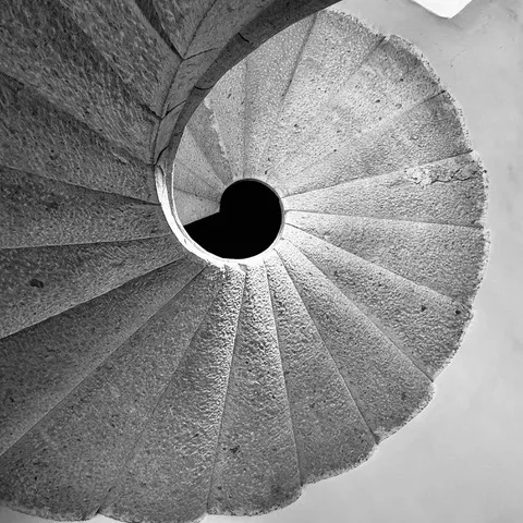 Scala a chiocciola in pietra vista dal basso, i gradini si avvolgono a spirale attorno al vuoto centrale, in bianco e nero