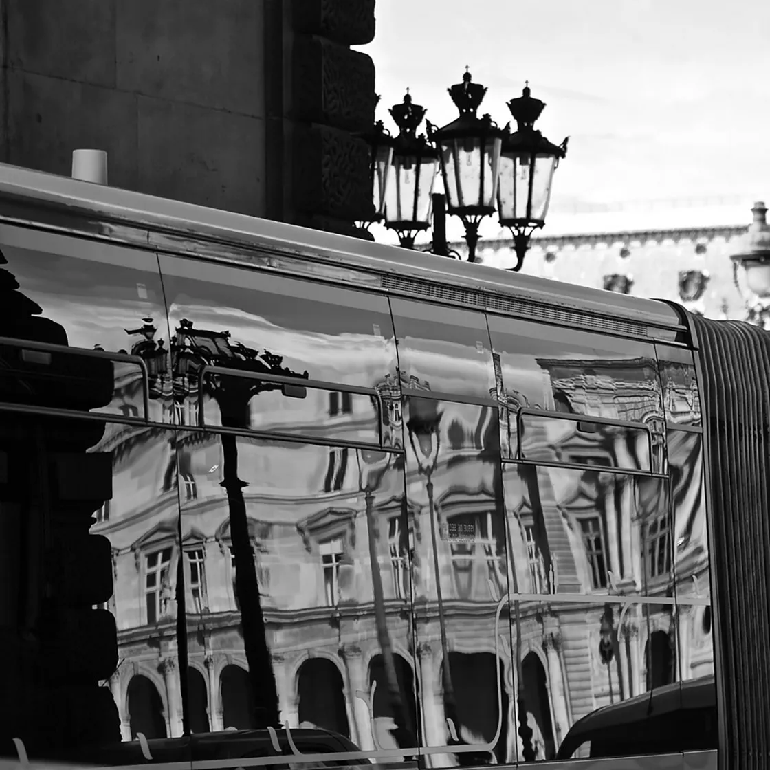 Facciata di un palazzo ottocentesco riflessa e deformata sulla fiancata lucida di un veicolo, lampioni ornati sopra, in bianco e nero