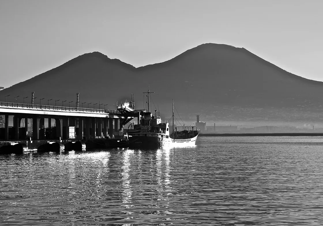 Una nave ormeggiata a un molo, con un monte dal profilo a due gobbe che si staglia nella foschia, in bianco e nero