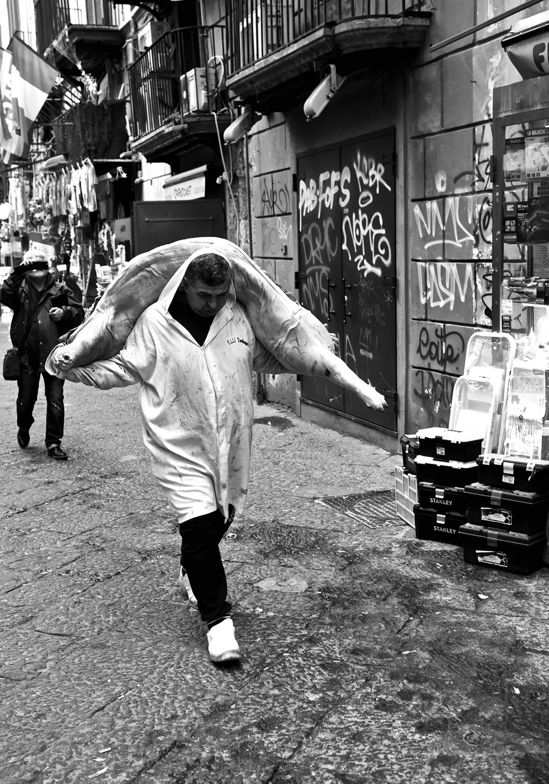 Un uomo in camice bianco cammina per un vicolo portando in spalla una carcassa, tra muri pieni di graffiti, in bianco e nero